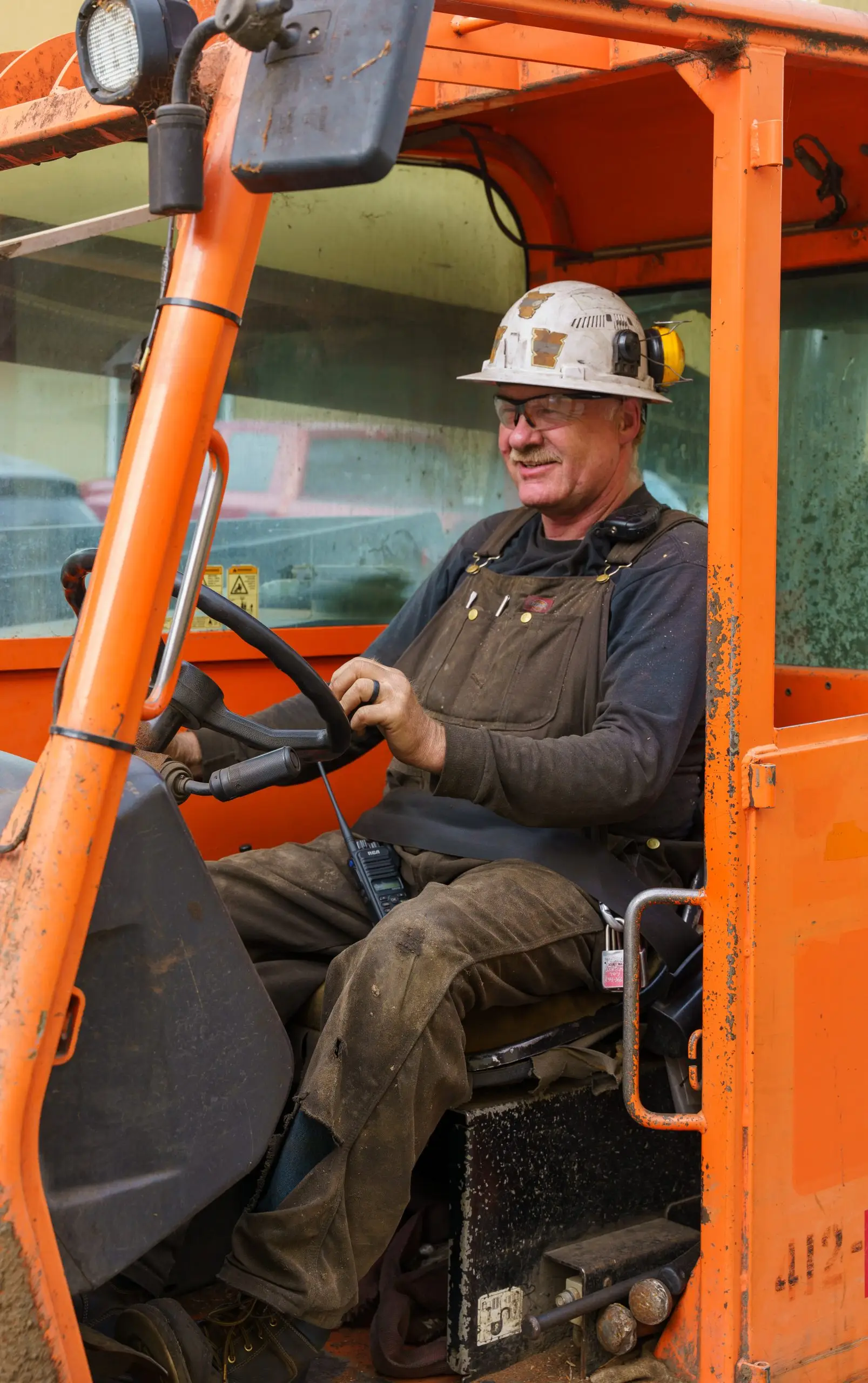 A loader operator at the controls in the cab, radio on his knee.