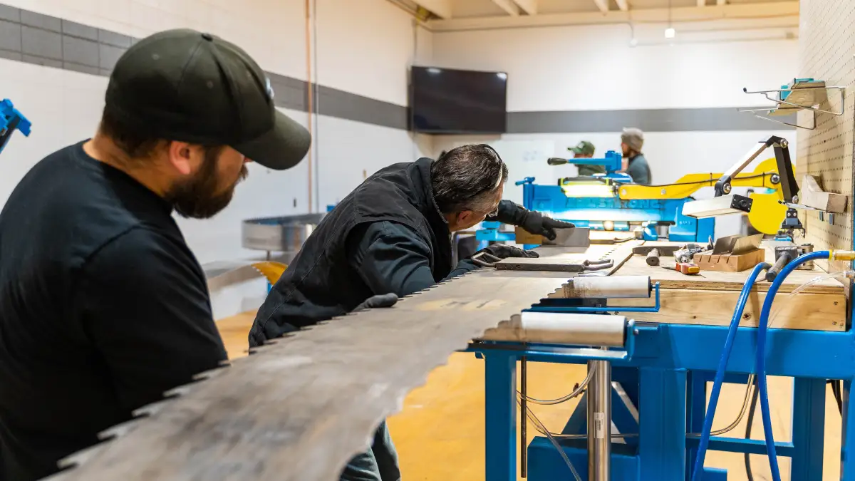 Two workers at the filing bench running the teeth of a wide bandsaw blade between them.