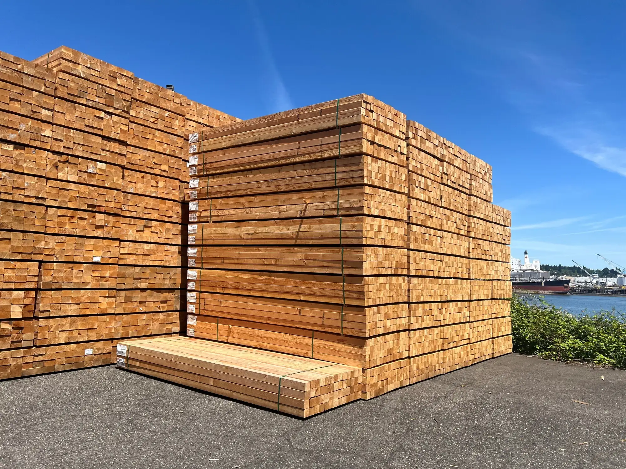 Banded and tagged packs of finished lumber stacked in the yard, with a ship at the dock on the waterfront behind them.